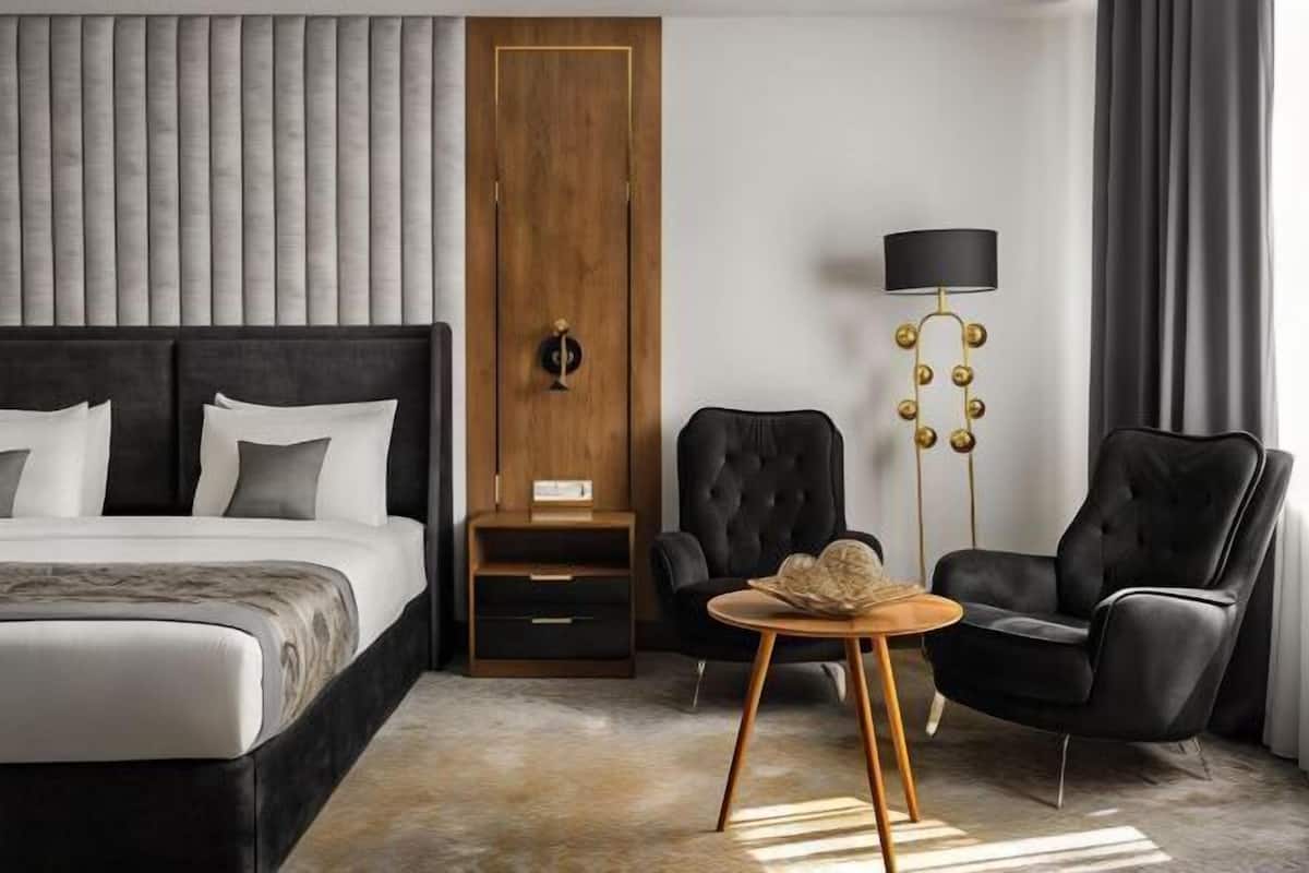 A modern hotel room with a large bed, a wooden wardrobe, a black armchair, a round wooden table, and a floor lamp.