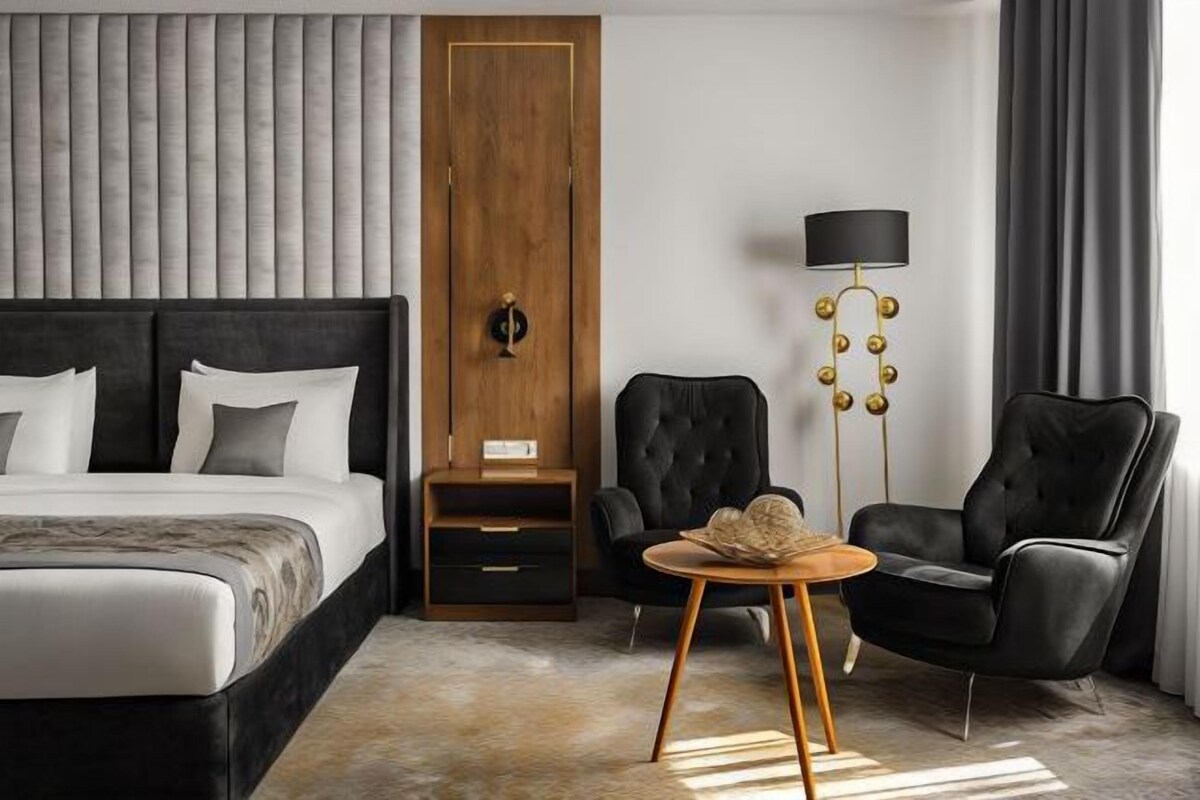 A modern hotel room with a large bed, a wooden wardrobe, a black armchair, a round wooden table, and a floor lamp.