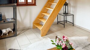 Une pièce avec un escalier en bois, une télévision, une petite table sur laquelle est posé un vase de fleurs et une fenêtre avec des stores.