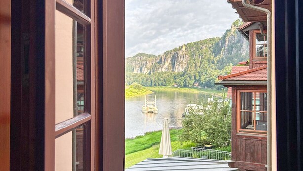 Une vue à travers une fenêtre ouverte, donnant sur un bâtiment à l’extérieur en bois, doté d’un balcon, et sur une rivière avec un pont en arrière-plan.