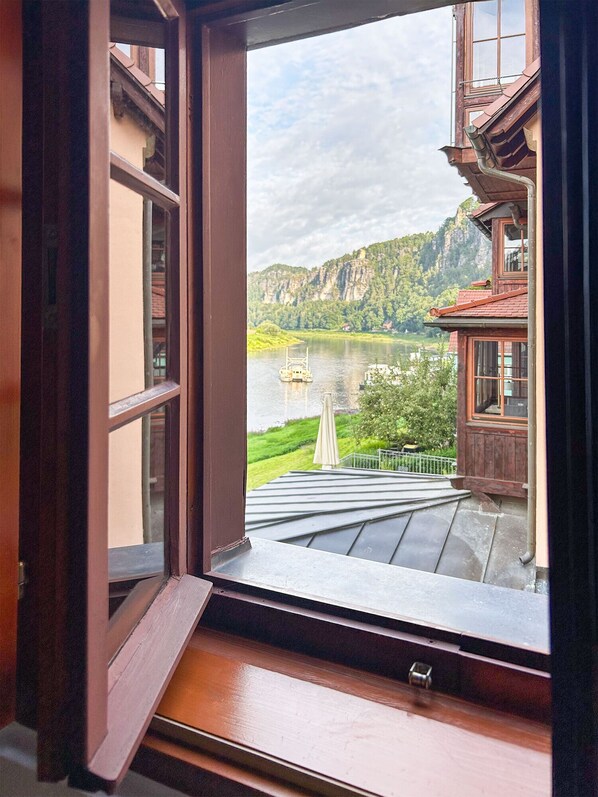 A view through an open window of a building with a wooden exterior, a balcony, and a river with a bridge in the background.