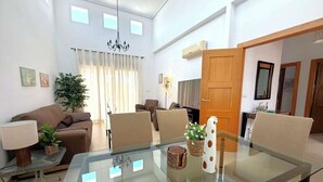 A modern living room with a glass dining table, sofas, a chandelier, and a large window with curtains. - Villa with private pool - CA13LT (La Torre Golf Resort)