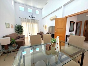 A modern living room with a glass dining table, sofas, a chandelier, and a large window with curtains.