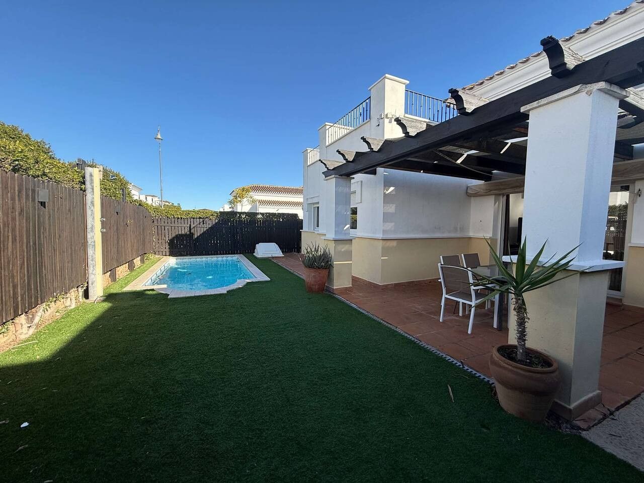 A backyard with a swimming pool, a patio with chairs, and a wooden fence.