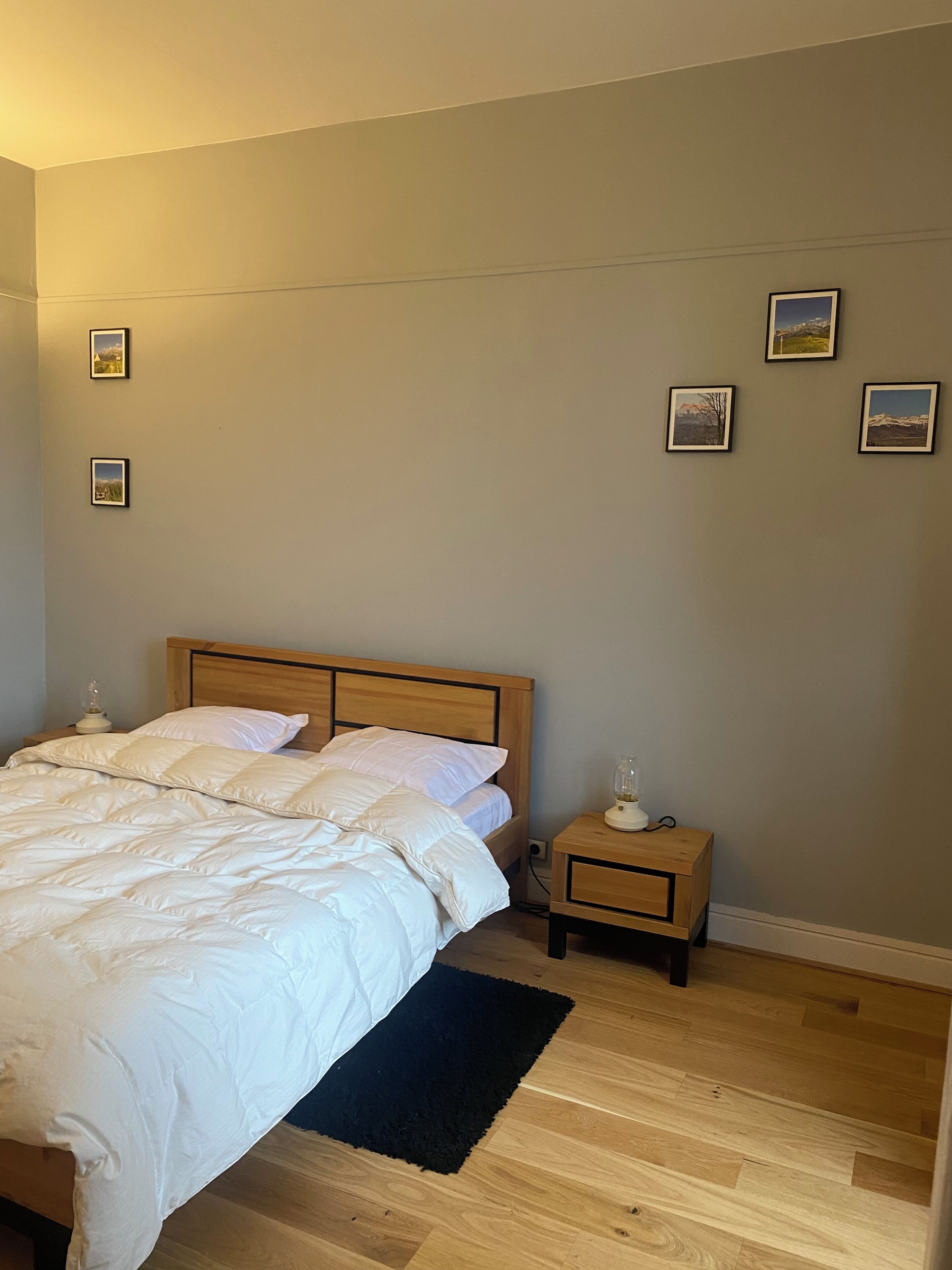 Quarto com cama de madeira, roupa de cama branca, tapete preto e mesa de cabeceira com abajur.