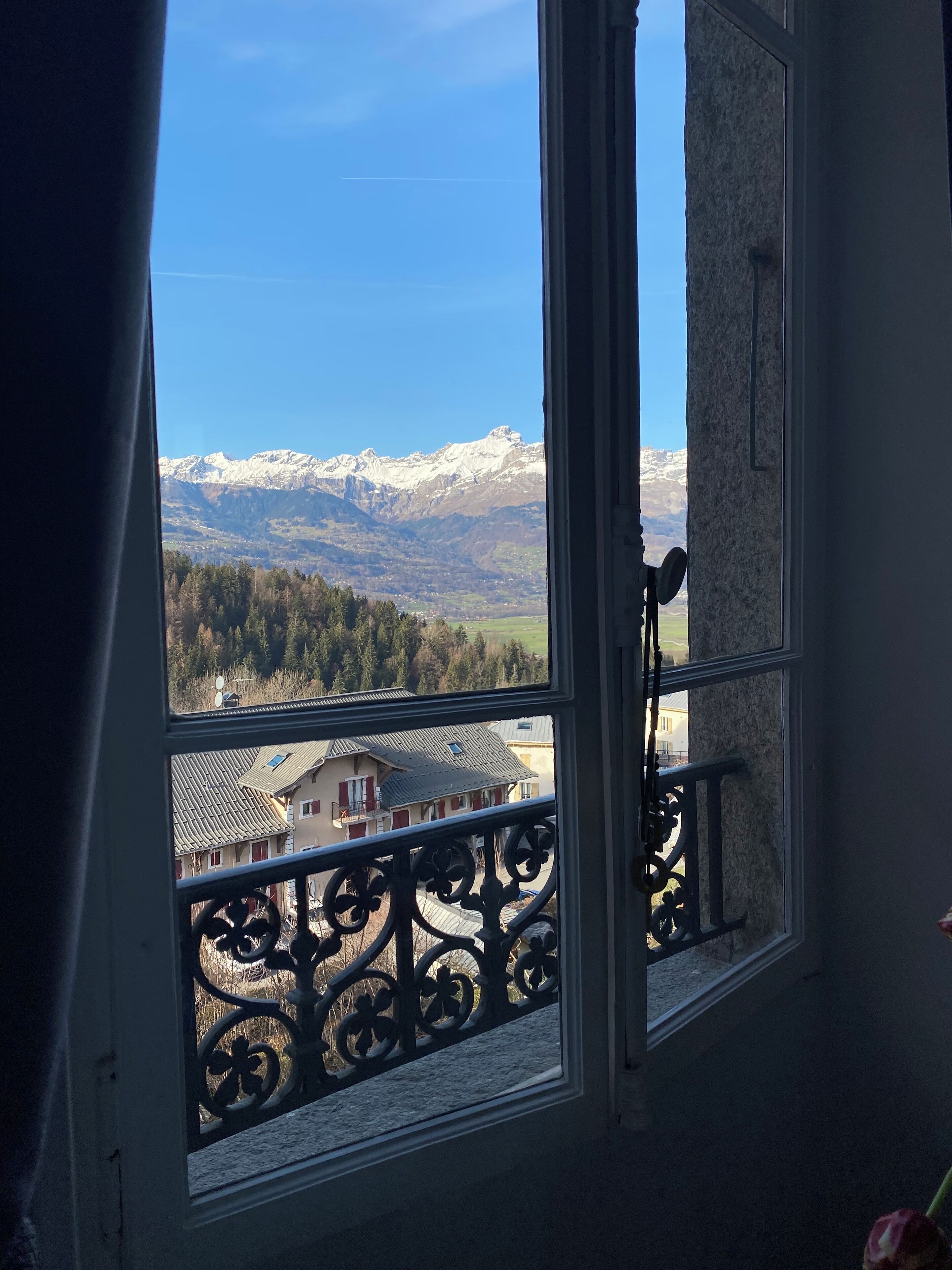 A window with a view of snow-capped mountains, a balcony with ornate metal railings, and a building with a sloped roof.