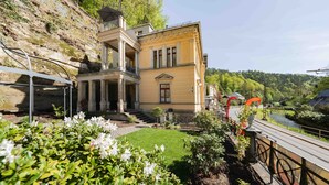 A historic building with a central entrance, a small garden, and a paved pathway leading to a bridge.