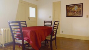 Un comedor con una mesa cubierta por un mantel rojo, rodeada de sillas de madera. Hay un radiador en el suelo a la izquierda y una puerta con un ventanal encima. Un cuadro está colgado en la pared a la derecha de la mesa.