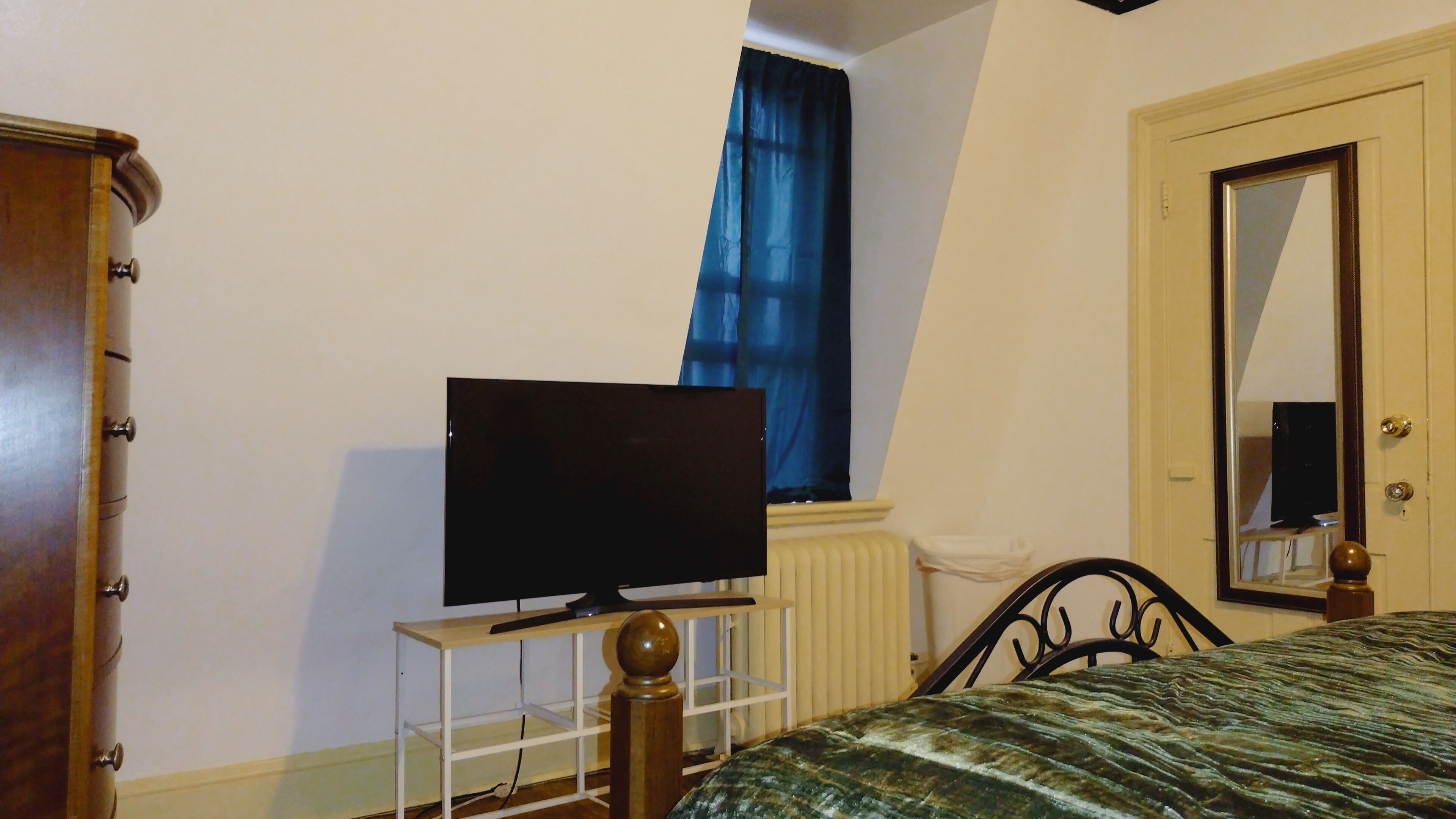 Un dormitorio con una cama, un televisor sobre un mueble, un armario, una ventana con cortinas azules y una puerta con un espejo.