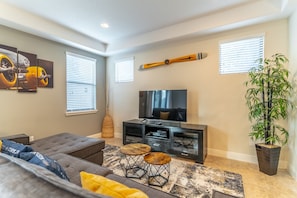 A living room with a large flat-screen TV, a grey sofa, two wooden stools, and a tall potted plant.