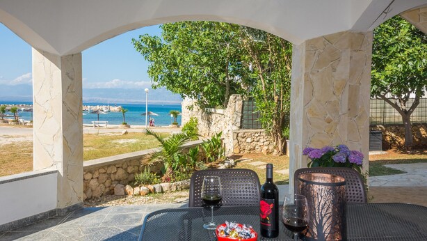 A table with a bottle of wine, a glass, and a bowl of snacks, set against a scenic view of the sea and a stone wall. - Apartments Andelic (Vir)
