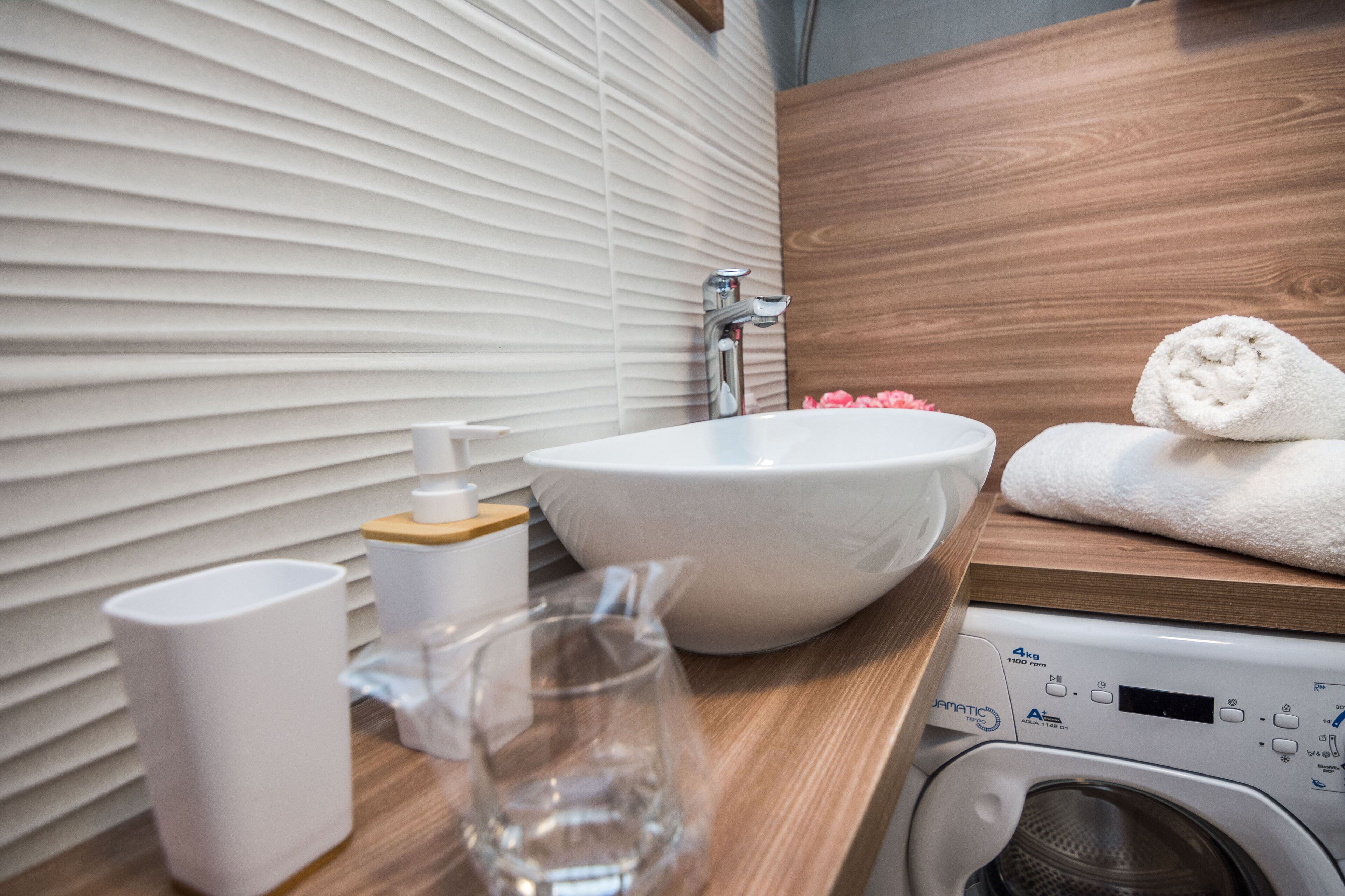 Ein Badezimmer mit einem weißen Waschbecken, einer Holz-Arbeitsplatte, einem weißen Handtuch, einer Glas-Karaffe und einer Waschmaschine.
