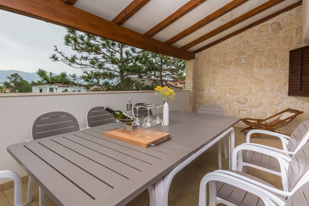 An outdoor patio with a table set for dining, surrounded by white chairs, and a stone wall in the background.