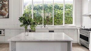 Una cocina moderna con una isla grande, muebles blancos, un ventanal con vistas a la vegetación y electrodomésticos de acero inoxidable.