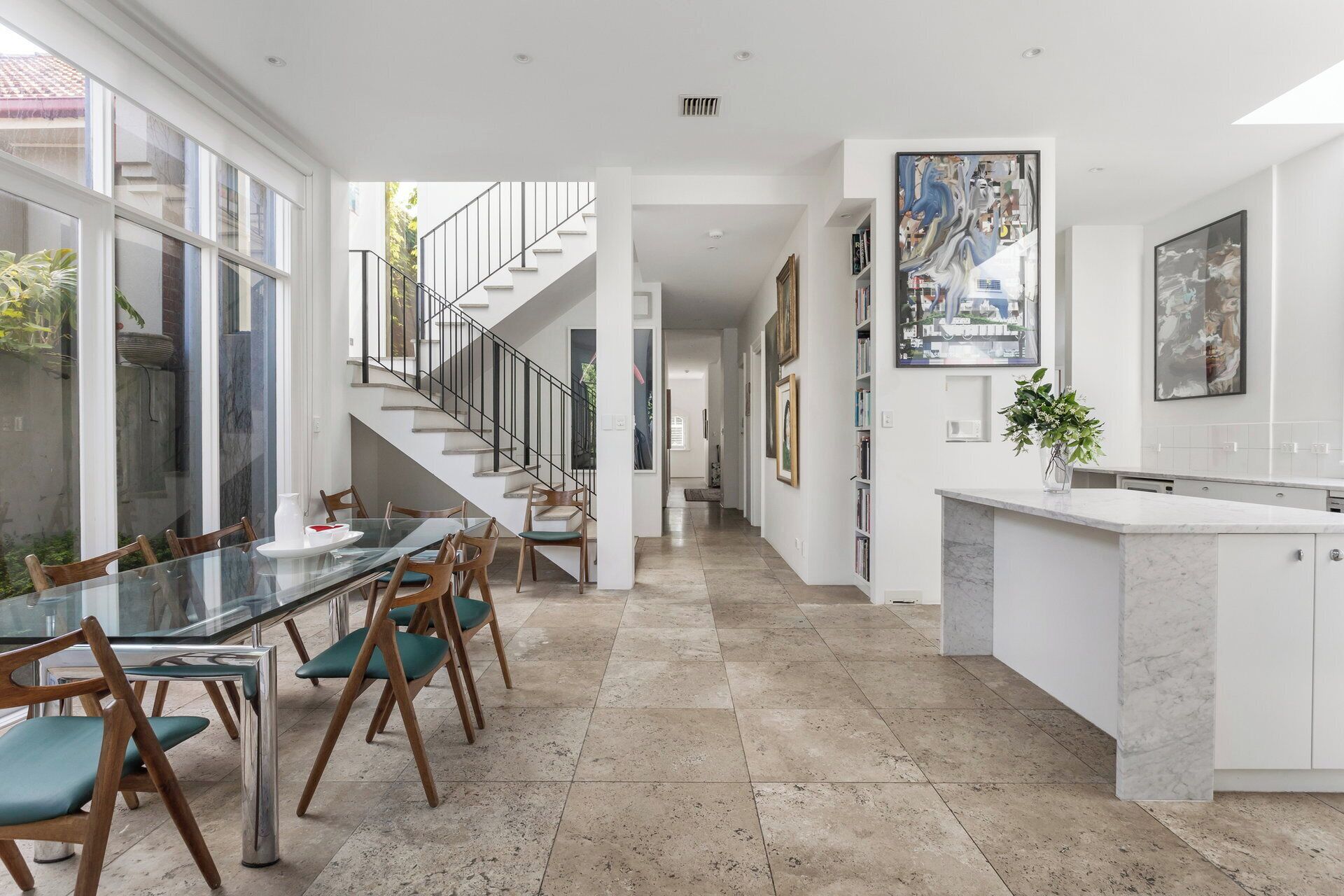 A modern kitchen with a marble countertop, a dining area with a glass table and wooden chairs, and a staircase leading to another level.