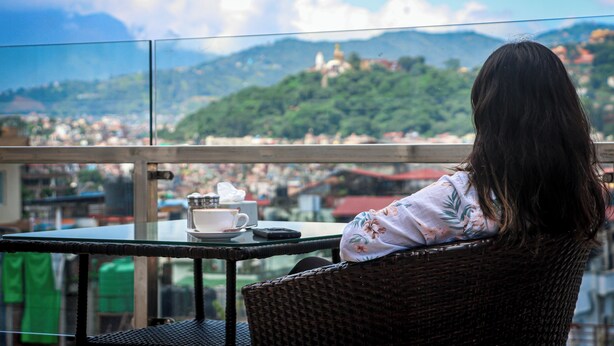 A person seated at a table with a cup and saucer, overlooking a cityscape with mountains in the background. - Hotel Simal (Kathmandu)