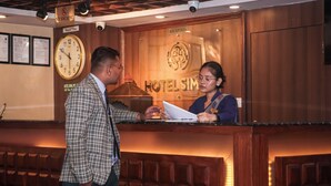 A hotel reception desk with a clock, a sign indicating no smoking, and a reception area with wooden paneling. - Hotel Simal (Kathmandu)