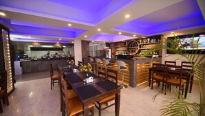 A restaurant with a long wooden table and chairs, a bar area with shelves of bottles, and a kitchen in the background. - Hotel Simal (Kathmandu)