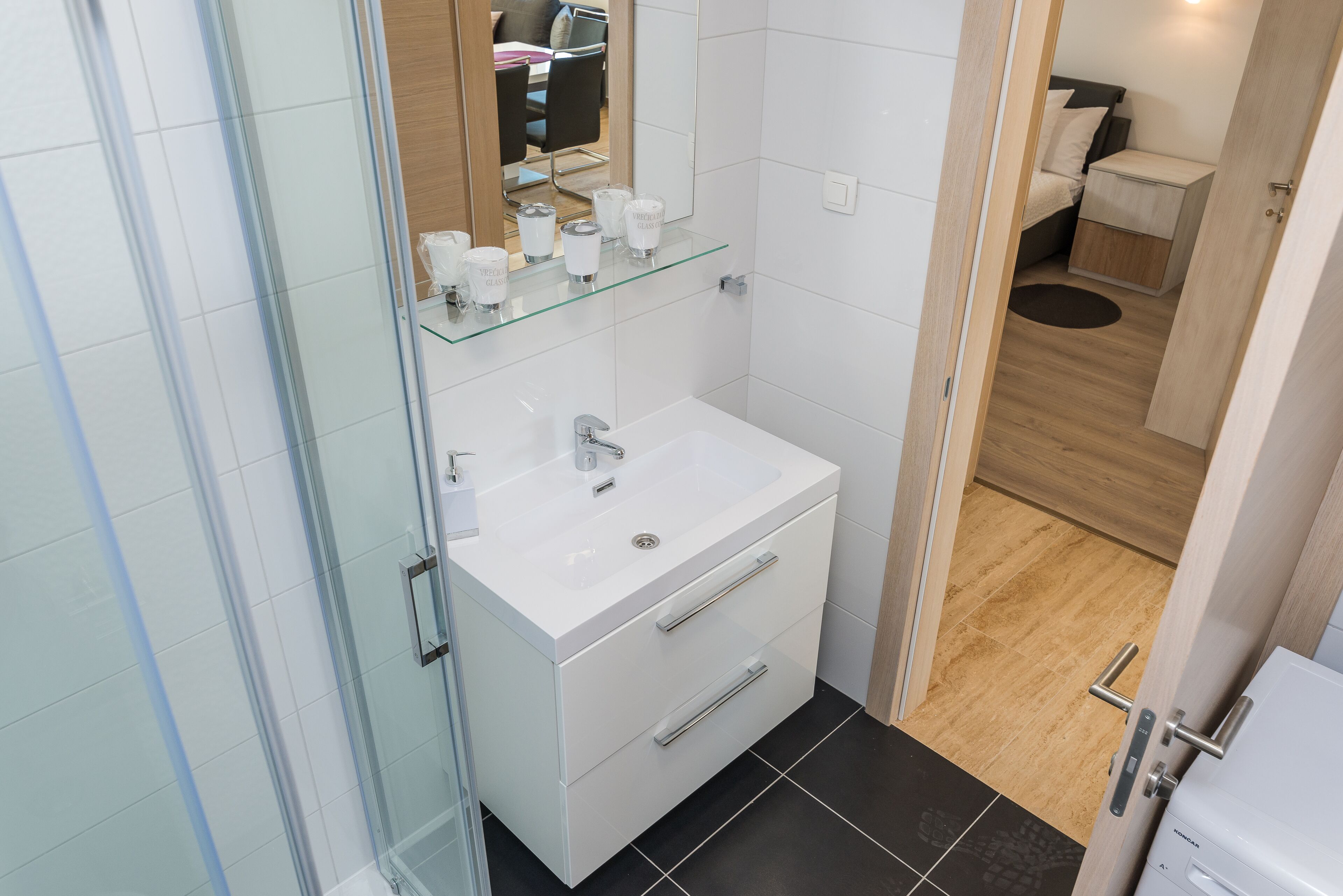 A compact bathroom with a glass shower enclosure, a white sink with a mirror above, and a small bedside table with a lamp.