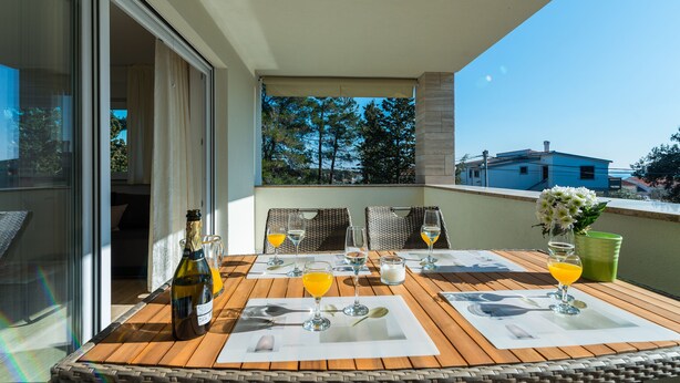 Uma mesa de madeira com uma garrafa de vinho, copos e um vaso de flores numa varanda com vista para árvores e edifícios.