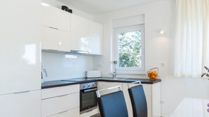 Cozinha moderna com armários brancos, bancada preta e janela com vista para árvores. Há duas cadeiras azuis e uma mesa branca.