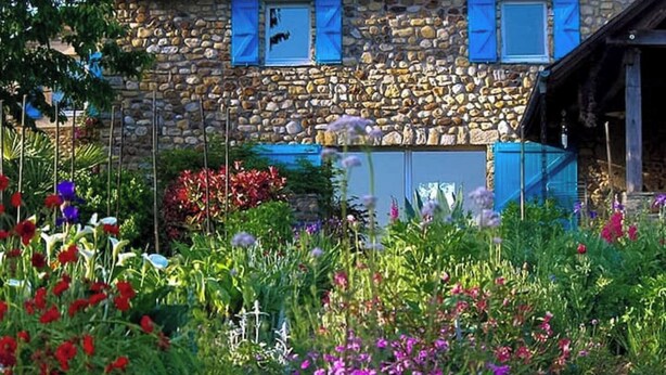 A stone building with blue shutters, surrounded by a garden with various flowers and plants. - GÎTE DE LA COLLINE AUX FRAMBOISES (Oloron-Sainte-Marie)