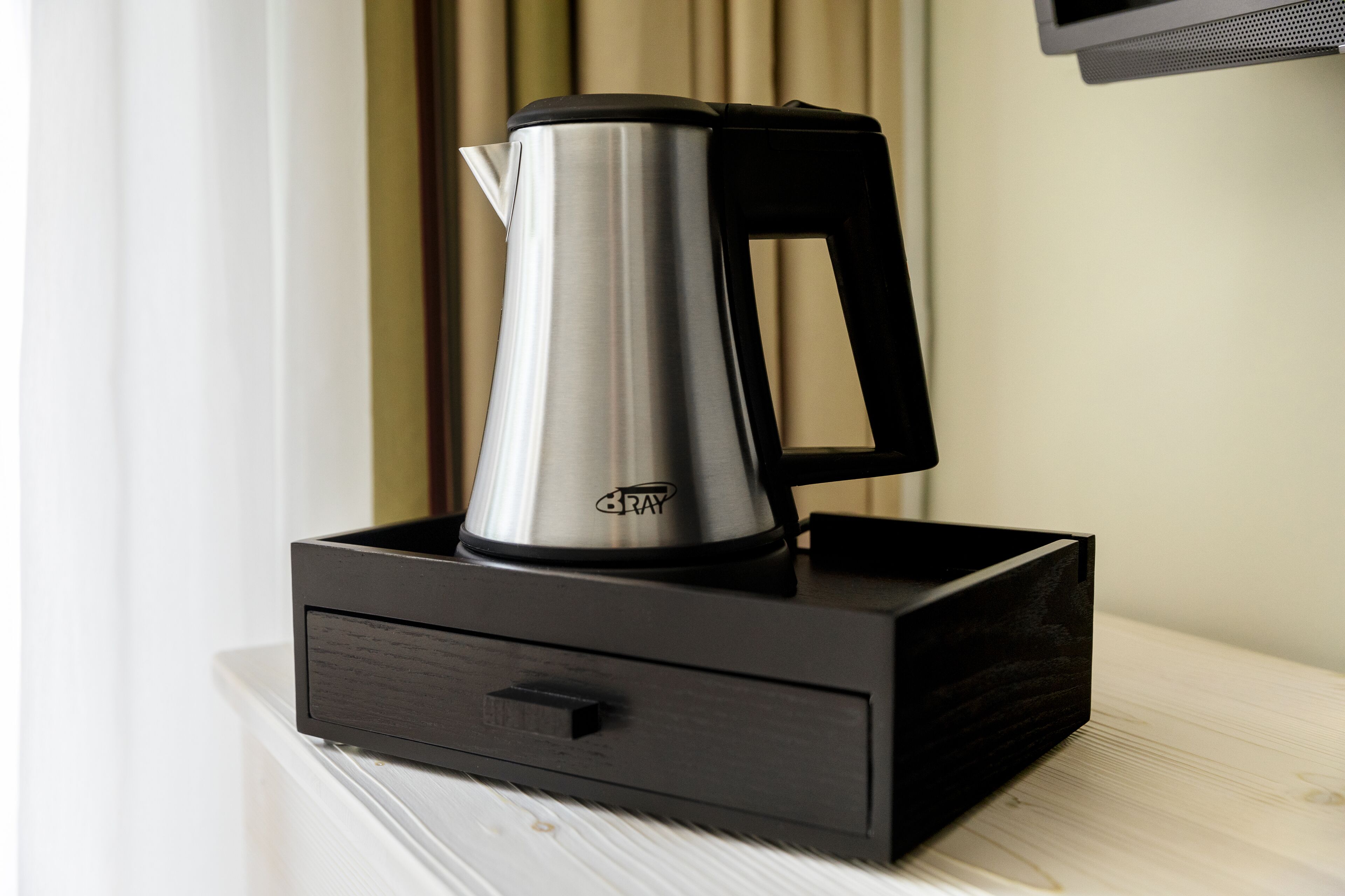 Une cafetière en acier inoxydable avec un manche noir, posée sur un plateau noir, elle-même placée sur une surface blanche.