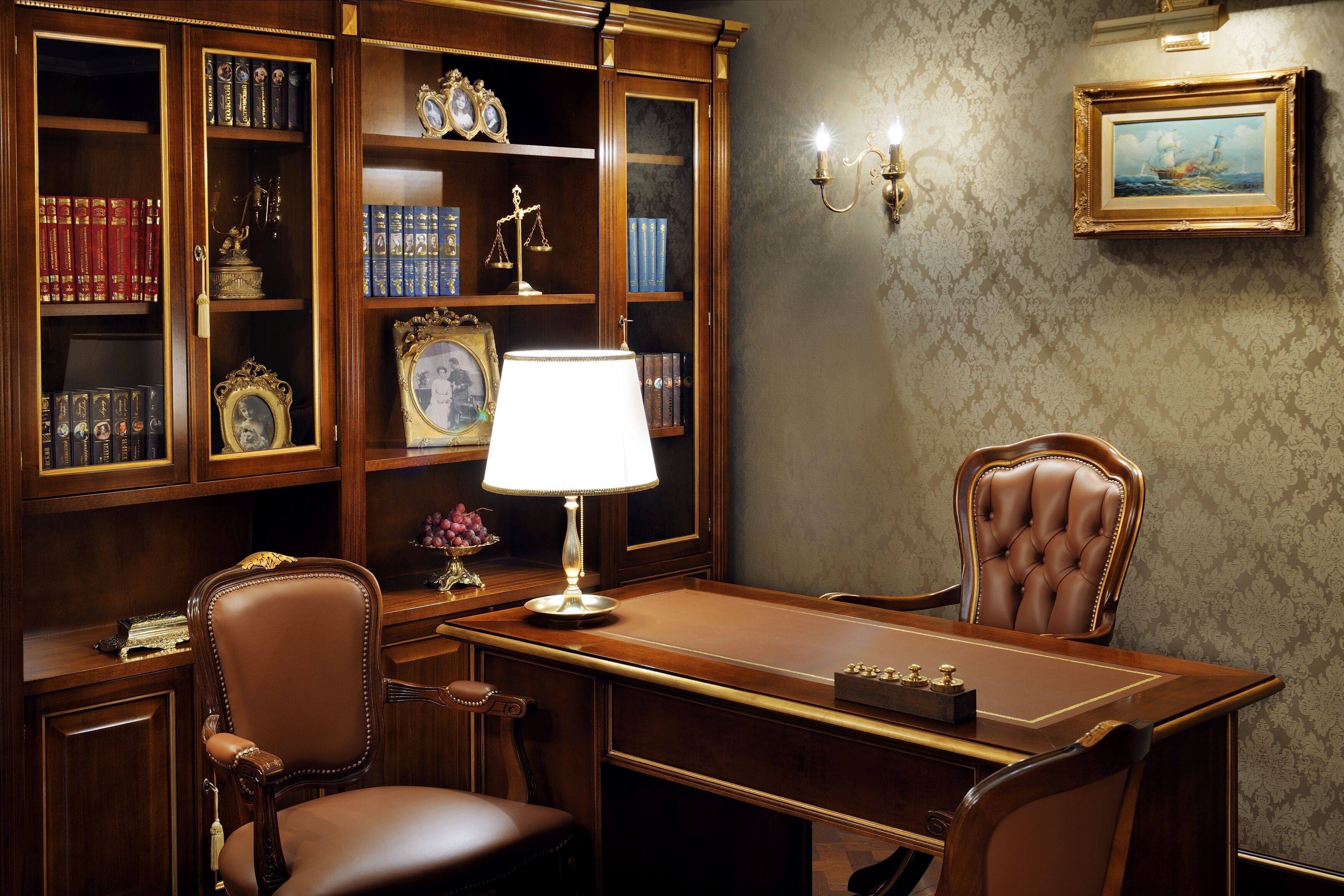 Een studeerkamer met een houten bureau, lederen stoelen, een lamp, boekenkasten vol boeken en decoratieve items, en een lijst met een afbeelding aan de muur.