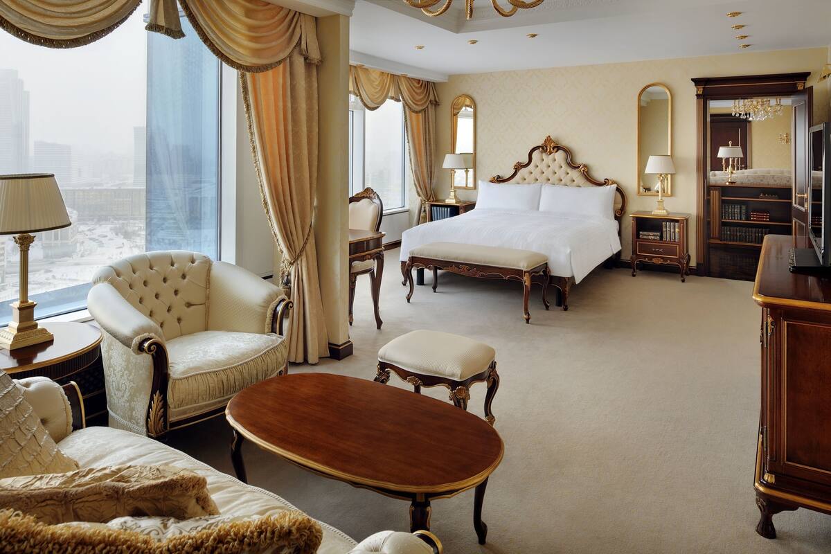 A luxurious hotel room with a large bed, a sitting area with a sofa and armchair, a wooden coffee table, and a view of the cityscape through large windows.
