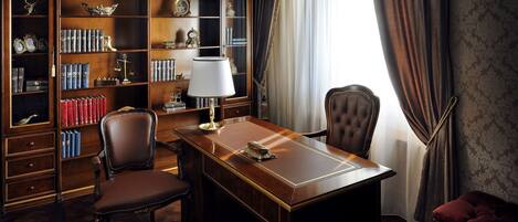 A study room with a wooden desk, leather chairs, a bookshelf filled with books and decorative items, and a large window with curtains.