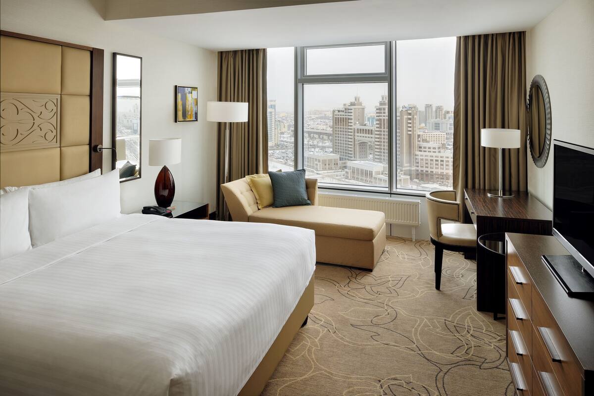A hotel room with a large bed, a sofa, a desk, a TV, and a view of the city through large windows.