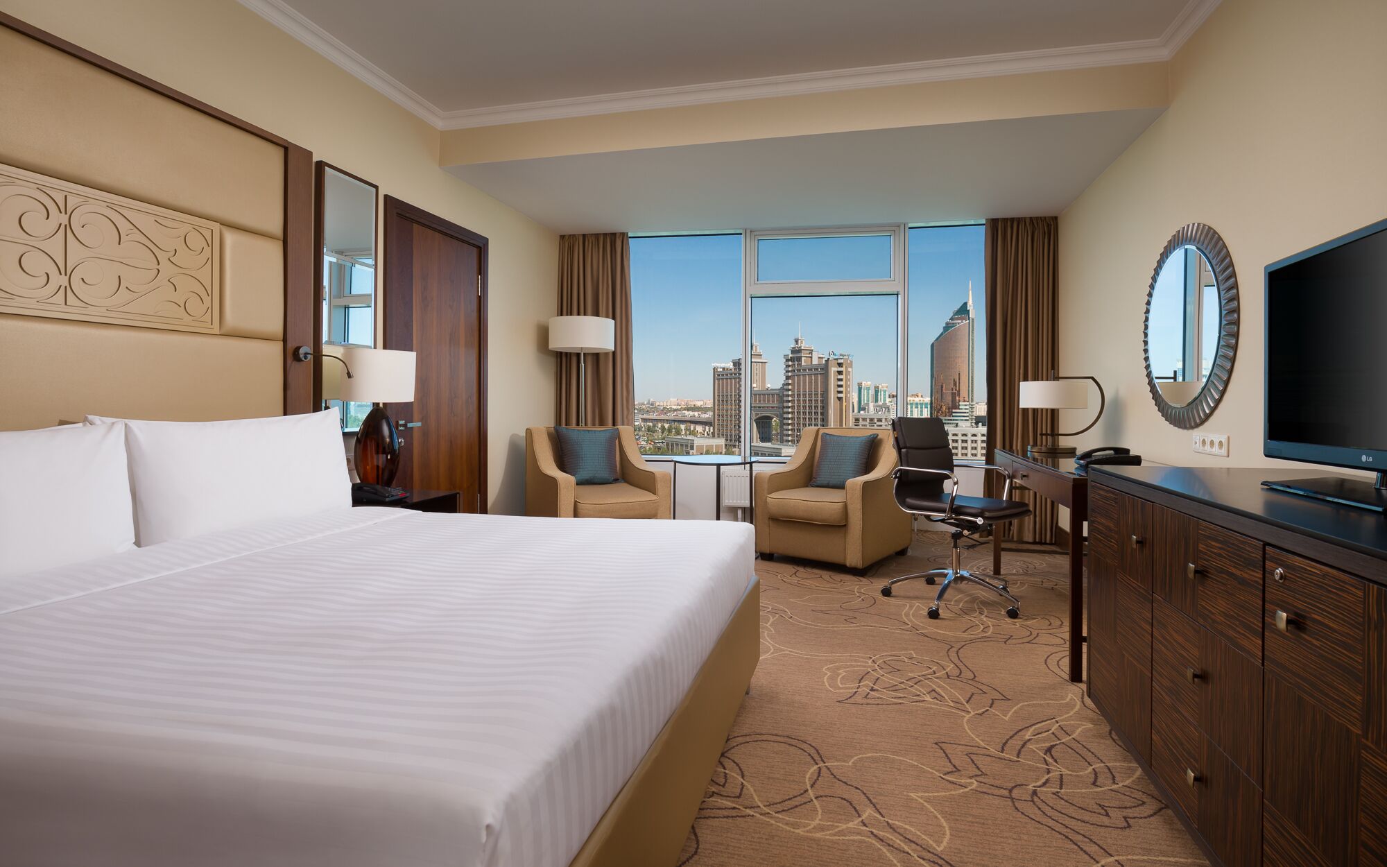 A hotel room with a large bed, two armchairs, a desk with a chair, a TV, and a view of the city skyline.