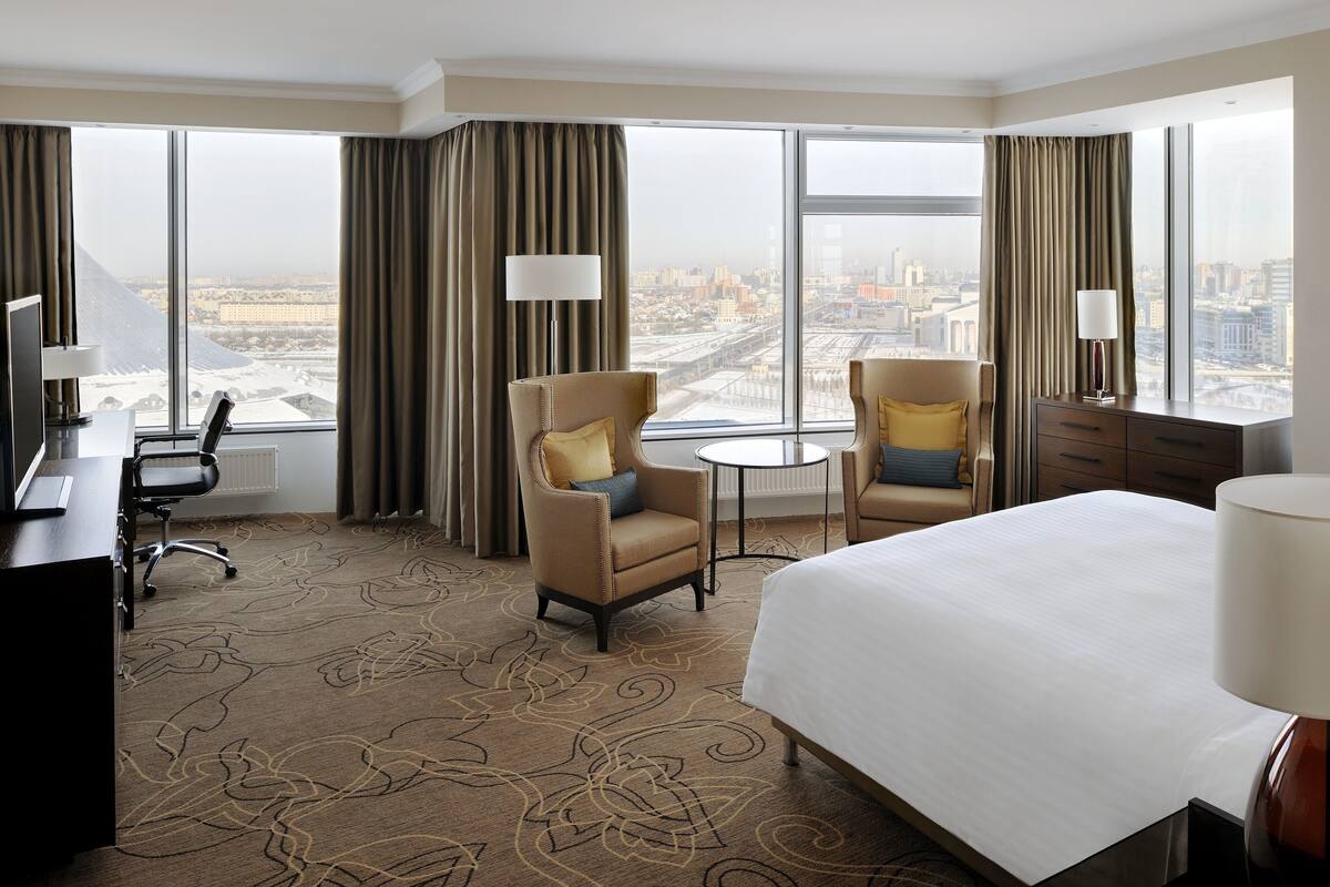 A hotel room with a large bed, two armchairs, a desk, and a view of the city through large windows.