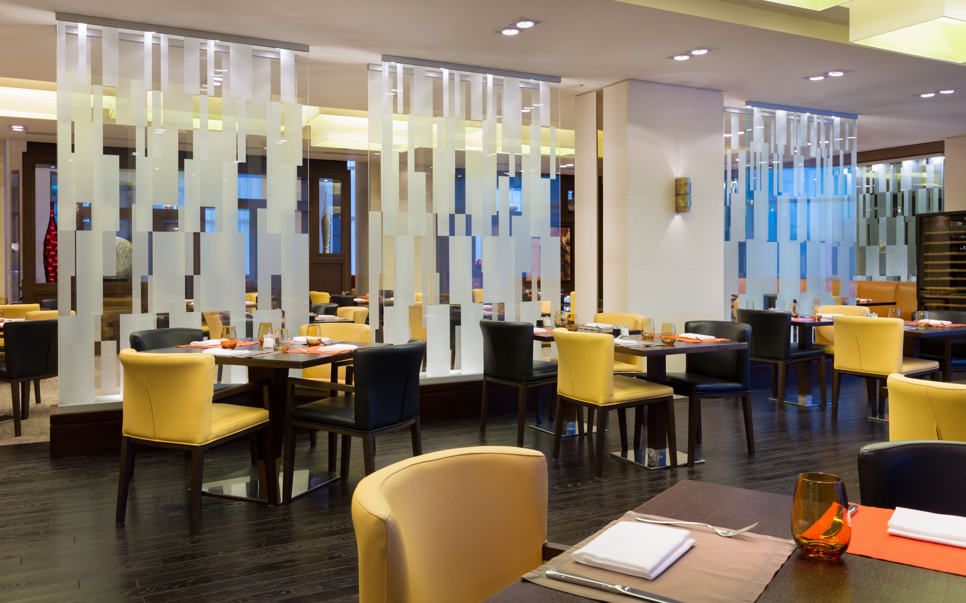 A modern restaurant interior with high tables, yellow and black chairs, and a decorative glass partition.