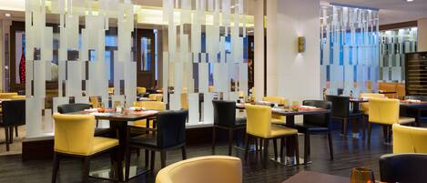 A modern restaurant interior with high tables, yellow and black chairs, and a decorative glass partition.