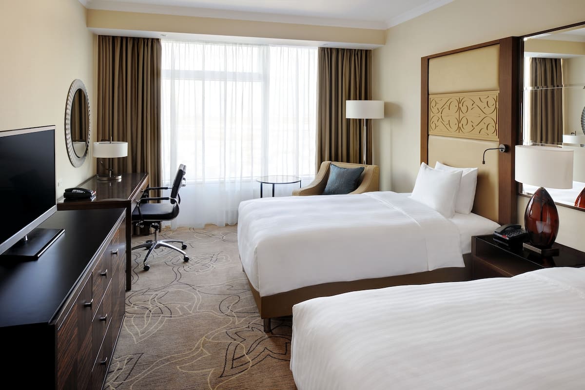 A hotel room with two beds, a desk with a TV, a chair, and a large window with curtains.