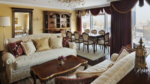A luxurious living room with a large sofa, a coffee table, and a chandelier. There is a dining area with a table and chairs, and a view of the city through large windows.