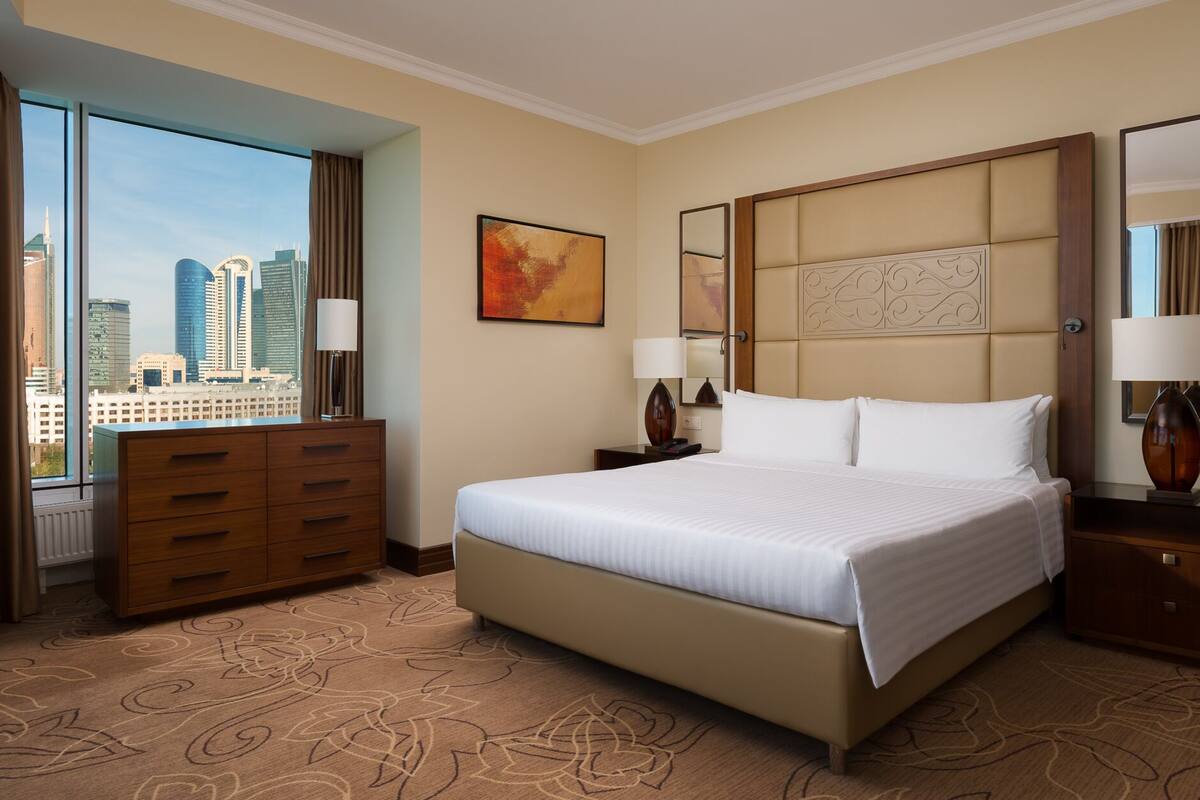 A hotel room with a large bed, a dresser, a nightstand, a mirror, and a view of the city skyline.