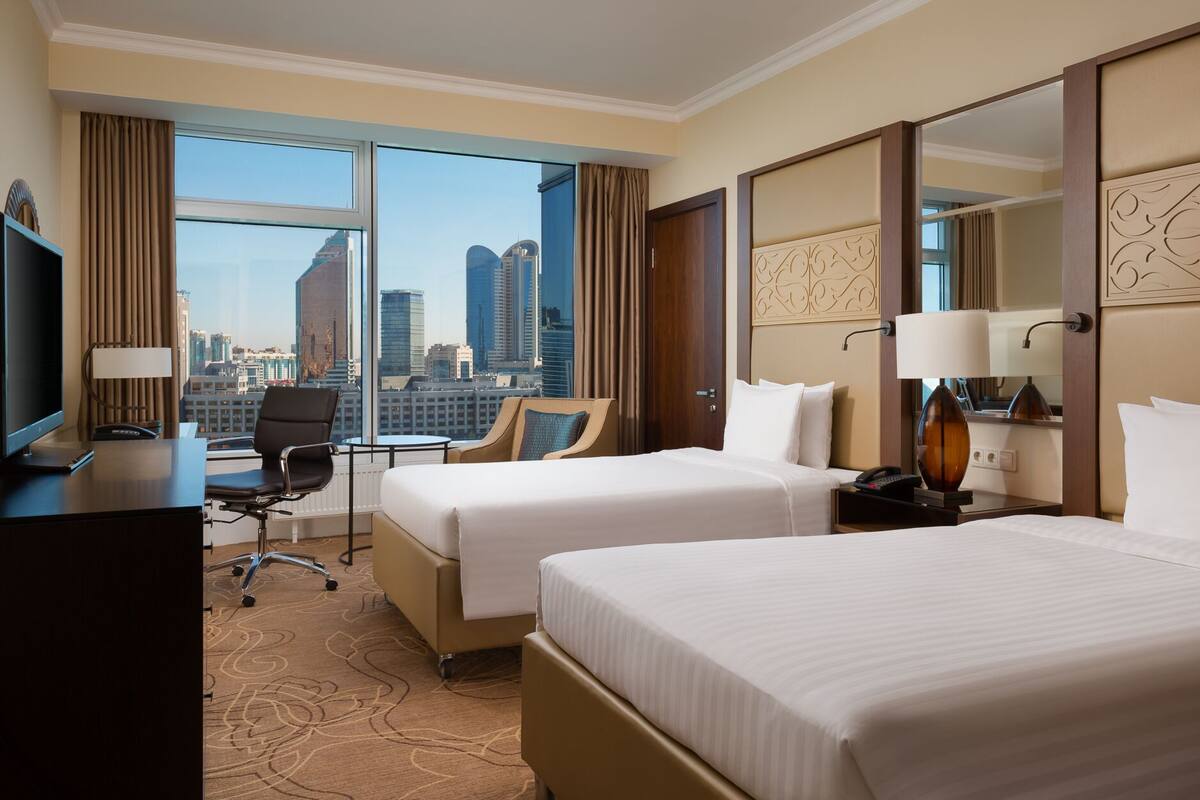 A hotel room with two beds, a desk with a computer, a chair, and a view of the cityscape.