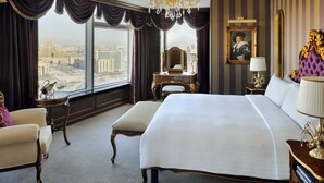Habitación de hotel amplia con una cama grande, un tocador con espejo, una silla y vistas a un paisaje urbano a través de ventanas amplias.