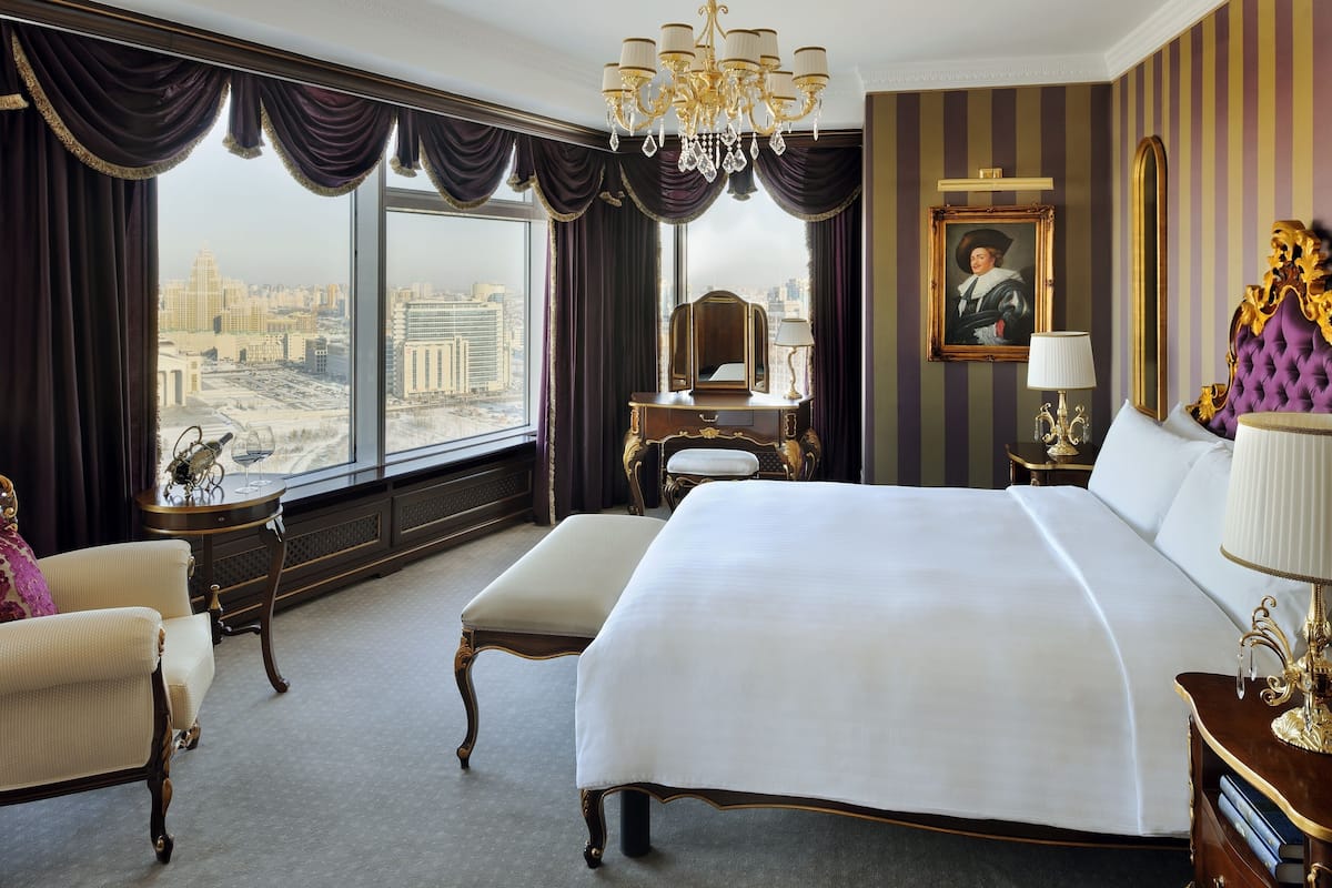 A spacious hotel room with a large bed, a vanity with a mirror, a chair, and a view of a cityscape through large windows.