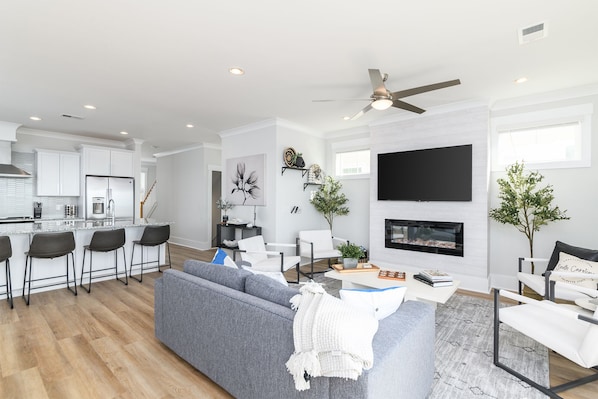 A modern living room with a grey sofa, a coffee table, and a fireplace. There is a kitchen area with white cabinets and a refrigerator.