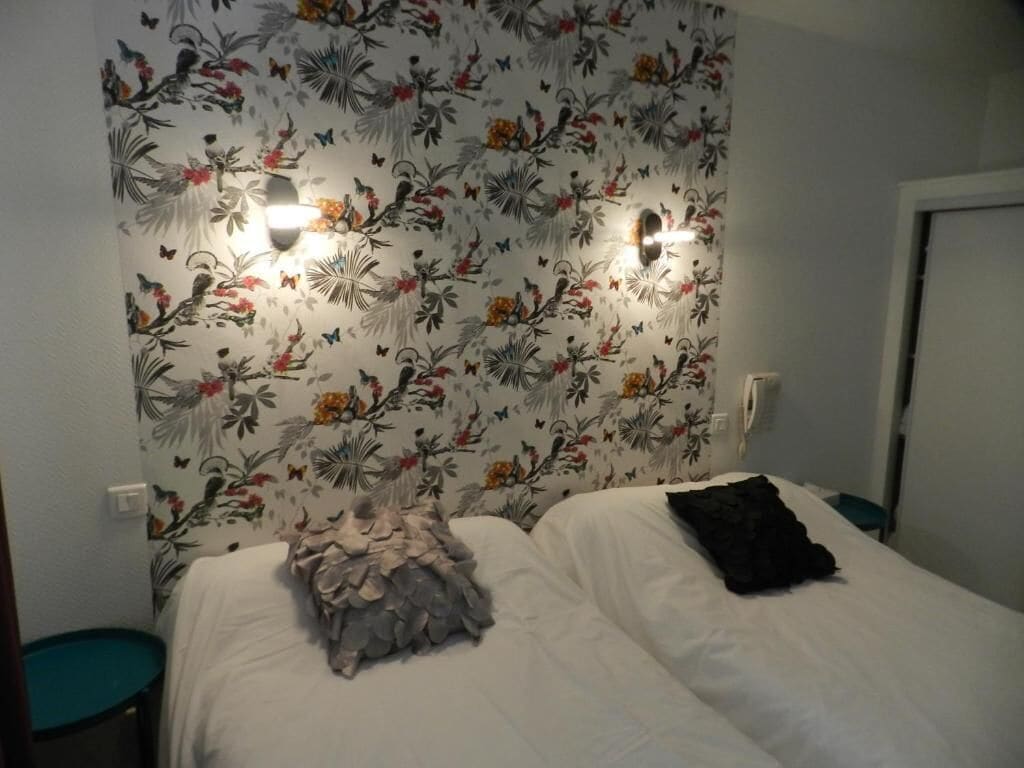Ein Schlafzimmer mit floralem Wanddesign, zwei Nachttischlampen, einem Bett mit weißer Bettwäsche und einem türkisfarbenen Sessel.