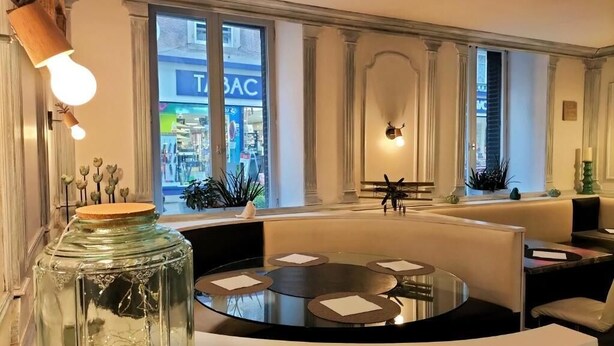 A modern interior with a round table, seating, and a view of a store through large windows. - HOTEL RESTAURANT HERARD  (Bourbonne-les-Bains)