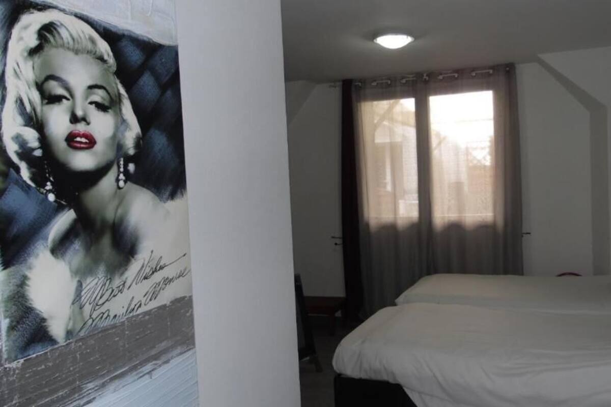 A hotel room with a large bed, two red pillows, a window with curtains, and a wall with a signed portrait.