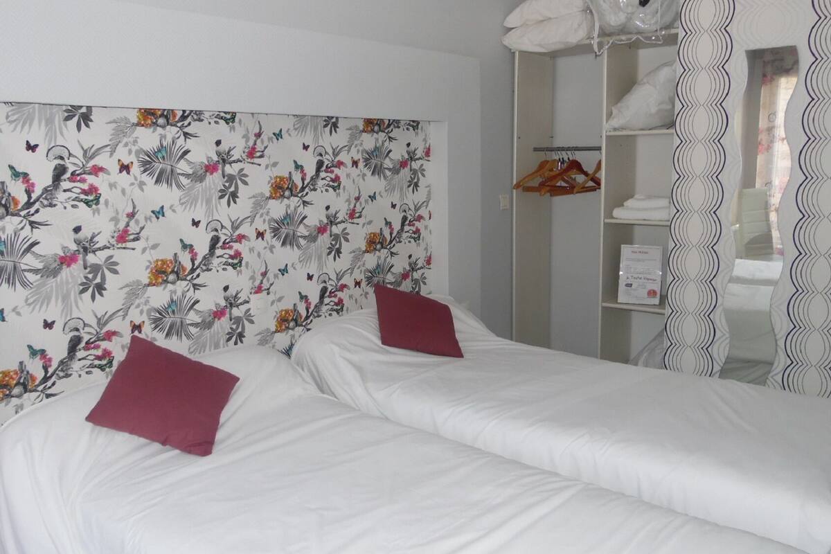 A double bed with white linens and two red pillows, a floral headboard, a wardrobe with hanging space, and a mirror with a wavy frame.
