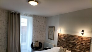 A hotel room with a large bed, a wooden headboard, a small round table with a chair, a window with curtains, and a wall-mounted light fixture. - HOTEL RESTAURANT HERARD  (Bourbonne-les-Bains)