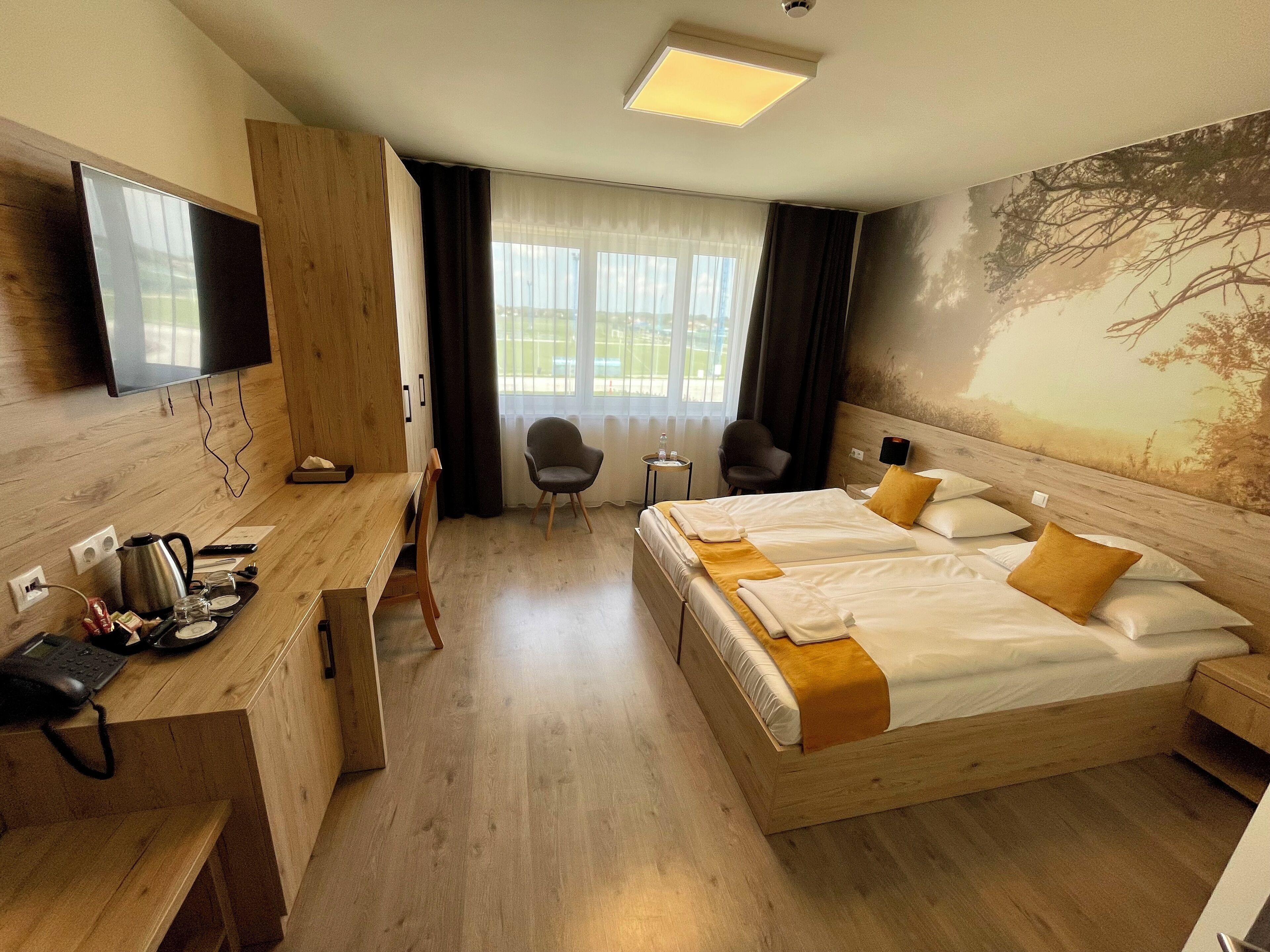 A hotel room with a wooden bed, a desk with a telephone and kettle, a chair, and a TV mounted on the wall.