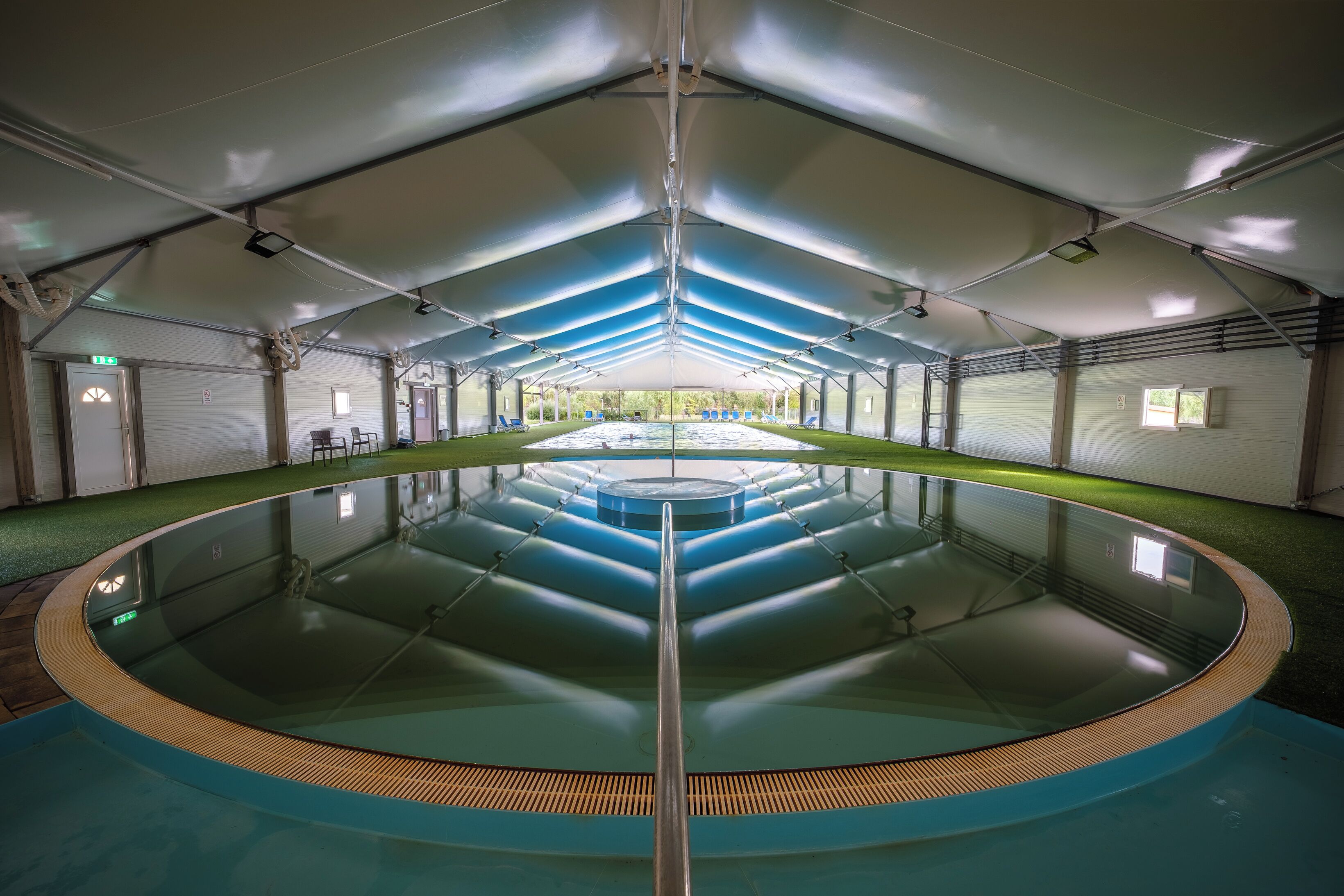 Uma grande piscina interior com uma plataforma de mergulho central, rodeada por espreguiçadeiras e uma área coberta com vista para o exterior.