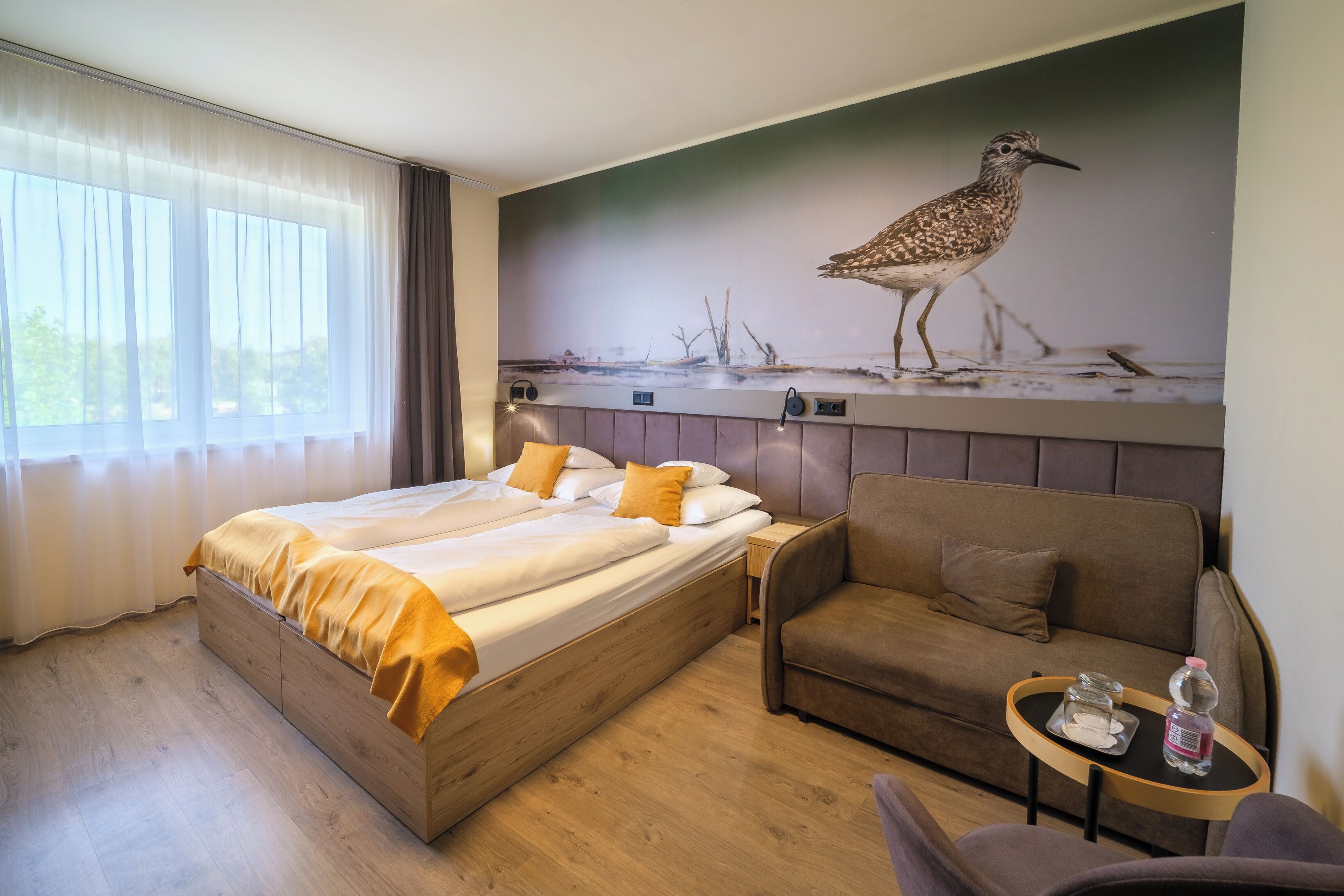 Habitación de hotel con cama, sofá, una mesita con una botella y un cuadro de un pájaro colgado en la pared.